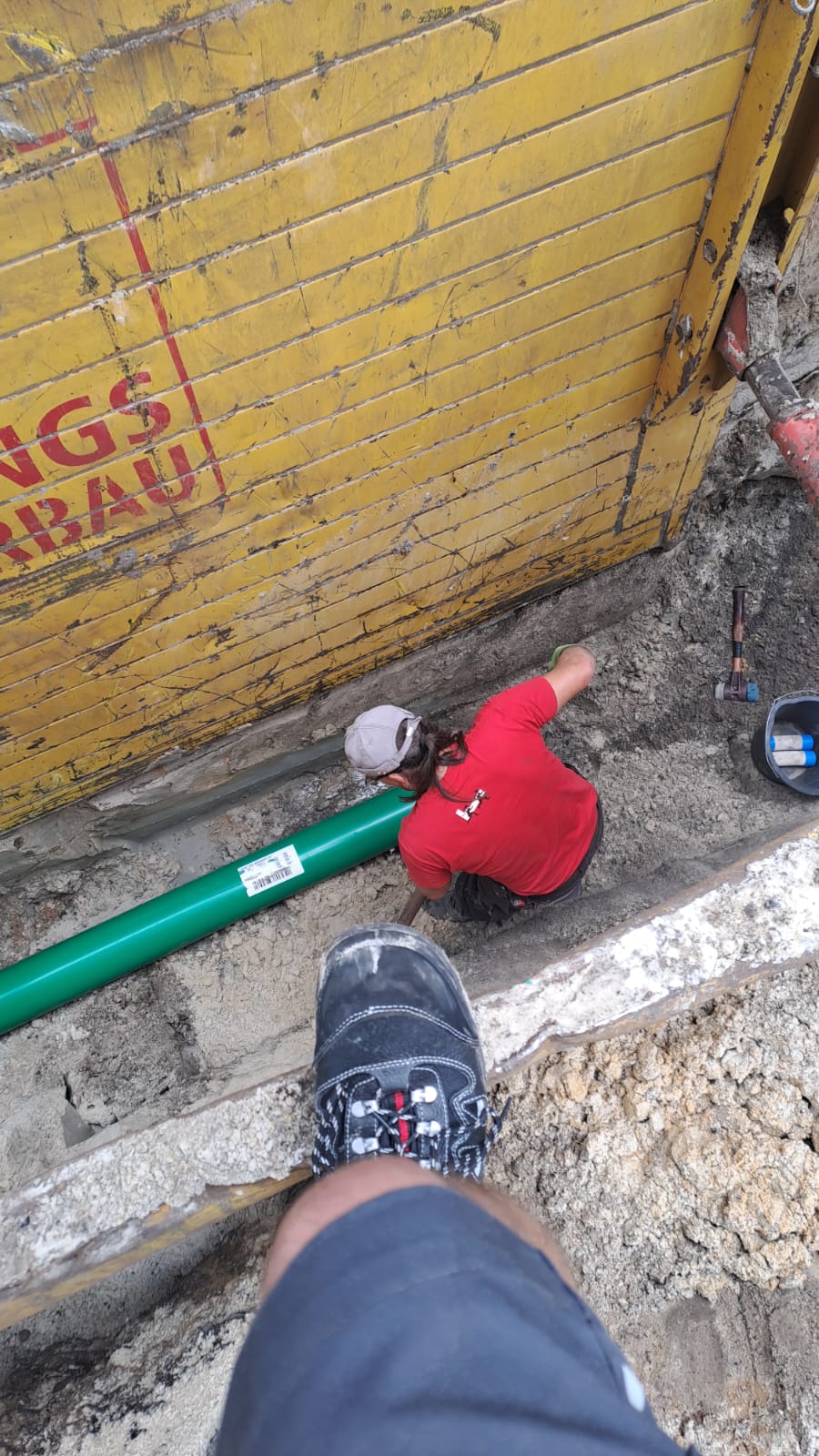 Arbeiter mit neutralem Blick in roter Jacke montiert grünes Rohr im Graben