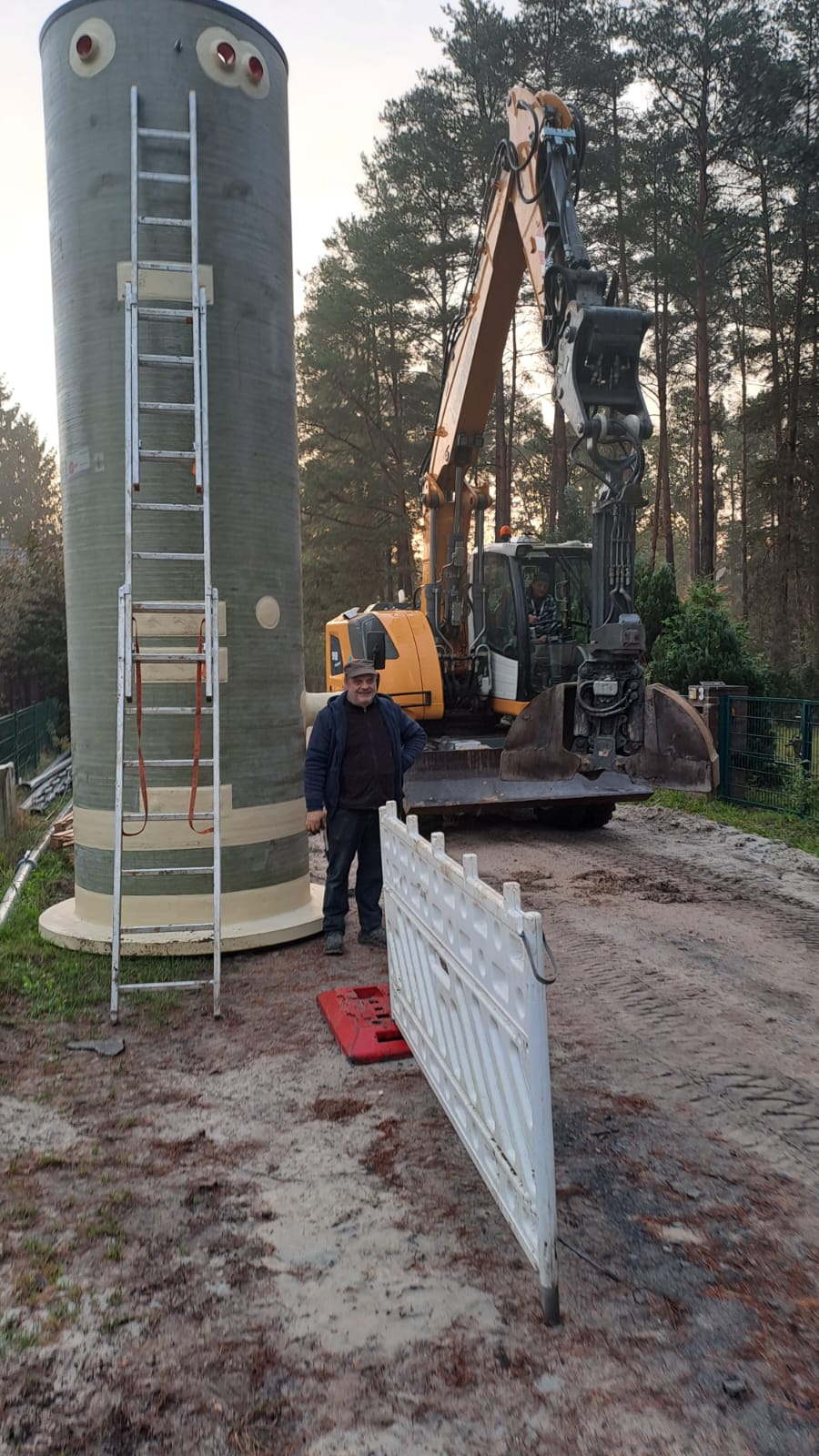 Mann laechelt vor grauem Tank gelber Baggergreifarm und Leiter im Wald